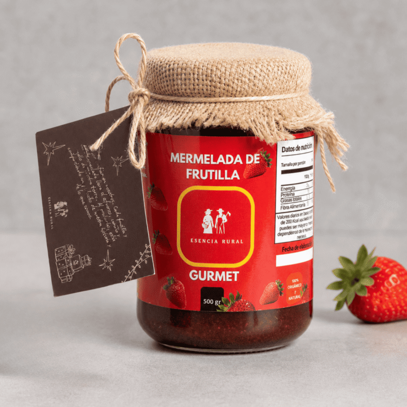 Frasco de vidrio de 500g de mermelada de frutilla gourmet marca Esencia Rural, con tapa cubierta de tela de arpillera rústica y etiqueta roja, acompañado de una frutilla fresca sobre fondo plomo.
