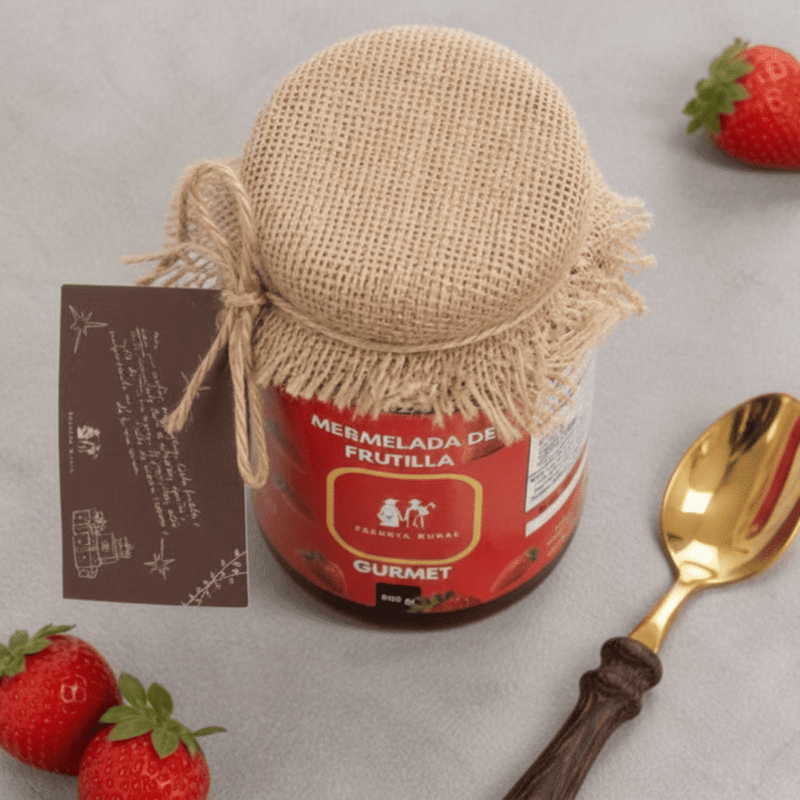 Vista superior de un frasco de mermelada orgánica Esencia Rural, destacando el empaque premium con tapa de yute amarrada con cordel, junto a una cuchara dorada elegante y frutillas frescas.